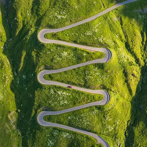 {
  "scene": {
    "description": "A winding mountain road switchbacks up a steep green hillside, seen from directly above by a drone",
    "season": "late spring, the hillside is covered in vivid green grass with scattered white wildflowers"
  },
  "composition": {
    "framing": "top-down, the road's switchback curves creating a zigzag pattern across the slope",
    "pattern": "the road is a thin grey line making 7 hairpin turns, each turn casting a tiny shadow on the hillside below it",
    "vehicle": "a single red car at the third hairpin, the only non-green element besides the road — it provides scale and narrative",
    "aspect_ratio": "4:5 vertical"
  },
  "camera": {
    "body": "DJI Mavic 3 Pro, Hasselblad camera",
    "focal_length": "70mm equivalent",
    "aperture": "f/5.6",
    "altitude": "600 feet",
    "iso": "100"
  },
  "lighting": {
    "time": "early morning, low sun from the east",
    "effect": "the switchback turns create alternating sun-lit and shadowed sections — each hairpin that faces east is bright, each that faces west is in its own shadow",
    "texture": "the morning light at this angle shows every contour of the hillside, the grass texture visible as a fine pattern"
  },
  "style": "Bernhard Lang's aerial road photography, the drone aesthetics of Daily Overview, infrastructure as graphic design",
  "skin_realism": "Highly realistic skin texture with visible pores, natural micro-imperfections, slight blemishes, no smoothing, no beauty filter, no airbrushed or plastic skin texture.",
  "negative_prompt": "airbrushed skin, plastic texture, waxy face, over-smoothing, beauty filter, CGI skin, straight road, flat terrain, urban, night, cloudy, multiple vehicles, traffic"
}