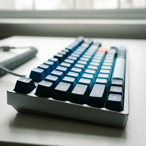 A mechanical keyboard sits on a clean white desk, and the photograph is about the keycaps — they are custom artisan keycaps in graduated blue tones, from dark navy at the Escape key to pale sky blue at the arrow keys, with a single orange accent on the Enter key. The shot is at a steep low angle, almost surface level with the desk, so the keycaps rise like a miniature cityscape of colored buildings. The nearest row of keys is in sharp focus, and the rows behind gradually soften into a smooth color gradient blur. Depth of field is impossibly thin — shot on a Sony A7R V with the Laowa 100mm f/2.8 2x Ultra Macro, creating a tilt-shift miniature effect without any digital processing. Single natural light from a window behind and above the keyboard, backlighting the keycap edges so each one has a bright rim against the shadow of the row behind it. The white desk surface goes slightly grey in the foreground shadow. Custom mechanical keyboard community photography, r/MechanicalKeyboards aesthetic, keycap collection hero shot, where the keyboard is not a tool but an art object. Highly realistic skin texture with visible pores, natural micro-imperfections, slight blemishes, no smoothing, no beauty filter, no airbrushed or plastic skin texture. Negative: airbrushed skin, plastic texture, waxy face, over-smoothing, beauty filter, CGI skin