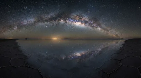 The Milky Way arching over a perfectly still salt flat, the crust surface so mirror-smooth that the entire sky — every star, the Milky Way band, the horizon glow — is reflected below as a perfect mirror image. A person would not know which half is sky and which is reflection. The salt crust shows hexagonal drying patterns near the edges. No moon — the Milky Way is the dominant light. 14mm, f/2.8, ISO 6400, 20-second exposure. Negative: light pollution, moon, people, text, watermark.