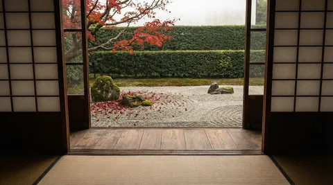 A Japanese zen temple interior, seen from the engawa veranda looking through open shoji screens at a karesansui dry rock garden. Tatami floor in the near ground, wooden threshold in the mid ground, raked gravel with circular patterns around three stones in the far ground, clipped hedge wall at the back. The shoji screen uprights create a frame-within-frame. Late autumn — a single red maple branch extends from the left, fallen leaves on the gravel. Soft overcast light. 45mm, f/8. Negative: people, bright sun, text, watermark.