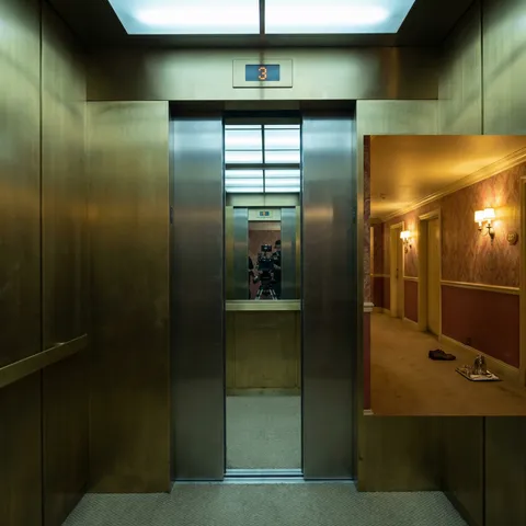 The camera is positioned inside a moving elevator as the doors open onto each floor of a building, revealing a different scene on each level — but this single frame captures the doors mid-open on one specific floor: a hotel corridor stretching into the distance, wallpaper in faded red and gold, a room service tray on the carpet outside room 237, a single shoe beside it (the second shoe is missing). The elevator interior is visible in the foreground — brushed brass panels, the floor indicator above the door showing '3', the mirrored back wall reflecting the camera itself (a deliberate fourth-wall acknowledgment). The corridor beyond the doors is lit by the yellow glow of period wall sconces, while the elevator interior is lit by a cooler overhead panel. The light temperature shift at the threshold — warm corridor, cool elevator — creates a visual boundary that the eye must cross. Shot on an ARRI Alexa Mini on a stabilized rig that would have been tracking into the corridor if this were video — but as a still, it captures the liminal moment of arrival, the doors not quite fully open, the corridor both inviting and warning. Zeiss Master Prime 21mm T1.3 at T2.0, 2.39:1. The Shining's Overlook Hotel corridor, Kubrick's one-point-perspective obsession.