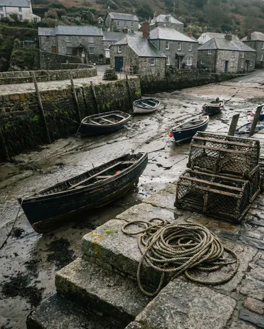 A fishing village harbor at low tide. Small wooden boats sit on mud and wet sand, tilted at angles. The harbor wall is stone, seaweed covering the lower section exposed by the tide. A coil of rope on the harbor wall. Lobster pots stacked near a stone stairway. The village rises behind — stone cottages with slate roofs. Overcast soft light. 35mm, f/8. Negative: tourists, modern boats, text, signage, watermark.