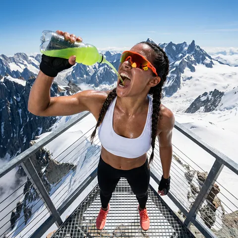 Dynamic overhead angle photograph of a muscular athletic woman with long braided hair and bright orange mirrored sport sunglasses, mouth wide open in a victorious yell, pouring a neon green sports drink from a translucent bottle into her mouth with the liquid stream clearly visible. She is wearing black fingerless training gloves. She's extremely sweaty with heavy perspiration covering her face, neck, shoulders and arms. Wearing a white fitted sports bra, black compression leggings, and bright coral running shoes with white soles. Shot from directly above at bird's eye view angle. Background: standing on the summit observation deck of a mountain with dramatic snow-covered peaks, blue sky, and wispy clouds visible below. Professional sports photography, photorealistic, high detail, dramatic alpine setting, intense post-workout summit celebration moment, 4K quality, sharp focus on subject with majestic mountain landscape surrounding.