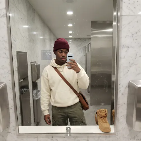 SHOT: Composition: Close-up mirror selfie front-facing of one visible subject in airport bathroom• Camera Settings: Smartphone camera wide-angle, auto exposure, natural lighting  • Film Grain: moderate grain  LENS EFFECTS:  • Optics: Slight fisheye distortion from bathroom mirror  • Artifacts: Minor reflections and water spots on mirror surface  • Depth of Field: Deep focus encompassing foreground and background  SUBJECT:  • Description: Young Black male, athletic build, curly dark hair under a burgundy wool beanie • Wardrobe: oversized cream colored hoodie with drawstrings, olive cargo pants, tan suede high-top sneakers with a brown leather crossbody messenger bag  • Grooming: Textured curly hair, gold chain necklace, manicured dark nails SCENE:  • Location: Airport bathroom with polished marble walls, stainless steel fixtures, tiled floor  • Time of Day: Indeterminate indoor lighting  • Environment: clean, modern airport setting with recessed LED lighting  VISUAL DETAILS:  • Action: Male subject holding smartphone angled for mirror selfie,   • Props: silver android phone  • Physics: Natural reflections and slight distortion in flat mirror  CINEMATOGRAPHY:  • Lighting: Bright LED recessed lights producing minimal shadows  • Tone: Travel casual with contemporary confidence  • Color Palette: Neutral whites, creams, olives, accented with gold accessories and burgundy tones  TEXT ELEMENTS:  • Visible Text: Partial sign on wall reading "Gate Information 12A"  • Typography: Standard airport directional signage font  • Placement: Sign mounted on marble wall behind subject  STYLE:  • Visual Aesthetic: Contemporary candid travel portrait