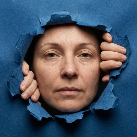 1) SUBJECT & IDENTITY
One adult woman, single subject.
Extreme close-up portrait, face occupies approximately 75–80% of the frame height.
Facial geometry, proportions, asymmetry, age cues preserved exactly as visible.
Natural skin texture with visible pores, uneven microrelief, and micro-shadows.
No beautification, no smoothing, no idealization.

2) SCENE
The woman's face is seen through torn deep blue paper.
The paper opening is irregular and asymmetric.
Background behind the subject remains dark and undefined.
No text, no graphics, no symbols.

3) POSE & PRESENCE
Head facing directly toward the camera.
Eyes looking straight into the lens.
Expression neutral and controlled.
Both hands grip the paper edges from behind.
Finger pressure visibly deforms the paper inward.
Paper tension increases near finger contact points.

4) CAMERA & COMPOSITION
Vertical framing.
Camera at eye level, perpendicular to the face.
No tilt, no rotation.
Lens behavior: short telephoto compression (approx. 85–100mm equivalent).
No wide-angle distortion.
Depth of field shallow but sufficient to keep face and paper edges in focus.

5) LIGHTING
Single frontal light source slightly above eye level.
Soft but directional light.
Shadows form under brow ridge, nose, lower lip, and within paper layers.
Light temperature neutral.
No cinematic contrast shaping.

6) WARDROBE & MATERIALS
Paper material (CRITICAL):
— Thick, multi-layered fibrous paper
— Visible cellulose fibers at torn edges
— Uneven fiber lengths protruding irregularly
— Layer separation visible where paper has torn
— Inner paper layers slightly darker due to density
— Matte surface with micro-roughness
— No glossy coating

7) MATERIAL BEHAVIOR (PAPER PHYSICS)
Paper tears follow fiber direction, not clean lines.
Edges show micro-fractures, feathering, and crushed fibers.
Paper bends inward under hand pressure.
Local creasing occurs near grip points.
Tear thickness varies along the edge.
No symmetry, no clean curves.
Paper holds partial rigidity but collapses locally where stressed.

8) BODY ↔ MATERIAL INTERACTION
Fingers compress paper edges.
Pressure points create localized indentations.
Paper edge thickness visibly increases where layers overlap.
Micro-shadows appear between paper layers and fibers.

9) COLOR & LIGHT INTERACTION
Deep blue paper color varies due to thickness and fiber density.
Darker blue in folded and layered areas.
Lighter blue on exposed fiber tips.
Skin tones remain neutral and unaffected by paper color bleed.
No color grading.

10) REALISM & OUTPUT
Photographic realism only.
Paper texture remains readable at close inspection.
Fibers remain visible under light.
No artificial grain, no HDR, no stylization.
No text, no logos, no watermarks.

11) AVOID
No poster aesthetics.
No cinematic mood language.
No smooth or "cut-out" paper edges.
No digital-looking tears.