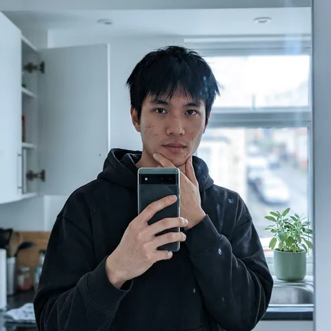 ```json
{
  "metadata": {
    "confidence_score": "high",
    "image_type": "photograph",
    "primary_purpose": "social media / private messaging"
  },
  "composition": {
    "rule_applied": "rule of thirds",
    "aspect_ratio": "9:16",
    "layout": "mirror selfie, single subject",
    "focal_points": [
      "Subject's face with subtle acne scars and intense dark eyes",
      "Phone in hand covering lower face slightly"
    ],
    "visual_hierarchy": "Eye goes straight to eyes and cheekbones in cool kitchen light, then to raised phone, finally to blurred window and background clutter.",
    "balance": "asymmetric, casual"
  },
  "color_profile": {
    "dominant_colors": [
      {
        "color": "Warm Olive",
        "hex": "#9CAF88",
        "percentage": "35%",
        "role": "skin tone and natural window light glow"
      },
      {
        "color": "Soft Charcoal",
        "hex": "#4A4A4A",
        "percentage": "25%",
        "role": "loose black hoodie and window shadow"
      },
      {
        "color": "Pale Cream",
        "hex": "#F9F7F4",
        "percentage": "20%",
        "role": "kitchen walls and countertop"
      },
      {
        "color": "Muted Sage",
        "hex": "#7A8B6F",
        "percentage": "10%",
        "role": "plant pot and small details"
      },
      {
        "color": "Deep Brown",
        "hex": "#3D2817",
        "percentage": "10%",
        "role": "hair and phone case"
      }
    ],
    "color_palette": "warm analogous",
    "temperature": "cool warm",
    "saturation": "low to moderate",
    "contrast": "soft"
  },
  "lighting": {
    "type": "indoor kitchen",
    "source_count": "window + overhead",
    "direction": "top and slight side",
    "directionality": "diffused through window glass",
    "quality": "soft, natural afternoon",
    "intensity": "moderate afternoon",
    "contrast_ratio": "low contrast",
    "mood": "intimate/casual",
    "shadows": {
      "type": "very soft",
      "density": "light",
      "placement": "gentle under cheekbones and jawline",
      "length": "none"
    },
    "highlights": {
      "treatment": "cool glow",
      "placement": "soft on temple, bridge of nose, cheekbones, hair edges, slight lens flare from window"
    },
    "ambient_fill": "strong cool",
    "light_temperature": "cool daylight from window"
  },
  "technical_specs": {
    "medium": "smartphone front camera mirror shot",
    "style": "WhatsApp-style casual selfie",
    "texture": "real skin texture, visible pores and minor blemishes",
    "sharpness": "sharp on face, slight motion blur on hand",
    "grain": "subtle phone noise",
    "depth_of_field": "shallow, background softly out of focus",
    "perspective": "straight on reflective surface, slight downward tilt"
  },
  "artistic_elements": {
    "genre": "private intimate selfie",
    "influences": [
      "WhatsApp mirror shots",
      "Afternoon casual vibe",
      "Real unfiltered moment"
    ],
    "mood": "soft/contemplative",
    "atmosphere": "Afternoon break between tasks, checking in with close friend",
    "visual_style": "lived-in kitchen, personal and unposed"
  },
  "typography": {
    "present": false
  },
  "subject_analysis": {
    "primary_subject": "Young man with subtle facial scars and straight black hair",
    "positioning": "center",
    "scale": "head and shoulders to mid-torso",
    "interaction": "taking mirror selfie with phone",
    "facial_expression": {
      "mouth": "relaxed, slight neutral expression",
      "smile_intensity": "minimal, thoughtful",
      "eyes": "direct intense gaze into mirror/camera, focused and present",
      "eyebrows": "natural, slightly furrowed",
      "overall_emotion": "quiet confidence, like sending photo to someone important",
      "authenticity": "completely candid private moment"
    },
    "hair": {
      "length": "short, just above collar",
      "cut": "straight natural cut",
      "texture": "slightly tousled from morning",
      "texture_quality": "undone strands, slightly flat at crown",
      "natural_imperfections": "uneven morning hair, visible hair line",
      "styling": "none, finger-combed only",
      "styling_detail": "pushed back casually",
      "part": "natural side",
      "volume": "textured roots, smooth ends"
    },
    "hands_and_gestures": {
      "left_hand": "holding phone up to mirror, fingers gripping sides",
      "right_hand": "lightly touching jaw near chin",
      "finger_positions": "relaxed grip on phone, fingers straight",
      "hand_tension": "none",
      "interaction": "casual selfie pose",
      "naturalness": "everyday motion"
    },
    "body_positioning": {
      "posture": "standing relaxed, slight lean toward reflective surface",
      "angle": "facing mirror straight",
      "weight_distribution": "even",
      "shoulders": "soft, both level"
    }
  },
  "background": {
    "setting_type": "small apartment kitchen",
    "spatial_depth": "shallow",
    "elements_detailed": [
      {
        "item": "Reflective Kitchen Mirror",
        "position": "entire background",
        "distance": "immediate",
        "size": "dominant",
        "condition": "clean",
        "specific_features": "slight dust, faint window reflection"
      },
      {
        "item": "White Kitchen Cabinet",
        "position": "left",
        "distance": "midground",
        "size": "medium",
        "condition": "everyday",
        "specific_features": "slightly open door"
      },
      {
        "item": "Small Plant on Counter",
        "position": "right on sink edge",
        "distance": "midground",
        "size": "small",
        "condition": "healthy",
        "specific_features": "sage pot, green leaves"
      },
      {
        "item": "Window Behind",
        "position": "top right",
        "distance": "background",
        "size": "medium",
        "condition": "clear",
        "specific_features": "blurred street view"
      }
    ],
    "wall_surface": {
      "material": "painted drywall",
      "color": "pale cream",
      "texture": "smooth with subtle imperfections"
    },
    "objects_catalog": "phone (Android-like), kitchen mirror, cabinet door, plant pot, window, water glass faintly visible",
    "background_treatment": "soft focus, natural haze"
  },
  "generation_parameters": {
    "prompts": [
      "Realistic smartphone mirror selfie of a young man with short straight black hair and subtle facial scars, wearing loose black hoodie, no beard, focused intense direct gaze into camera with quiet confidence. One hand holding phone steady, other hand lightly touching jaw. Cool natural window light, clean kitchen mirror background with subtle window reflection, white cabinet door on left, small sage plant pot on counter edge, blurred window view. Authentic private WhatsApp-style photo, natural skin texture and imperfections, shallow depth of field, contemplative afternoon moment.",
      "Candid kitchen mirror shot of young man with black hair in casual hoodie, natural window lighting, sending selfie to someone close, hyper-realistic smartphone photo with real details."
    ],
    "keywords": [
      "mirror selfie",
      "afternoon light",
      "subtle scars",
      "straight black hair",
      "casual hoodie",
      "kitchen mirror",
      "whatsapp vibe",
      "intimate moment",
      "soft window light",
      "authentic no filter"
    ],
    "technical_settings": "Android front camera, natural pose, cool window light, ISO 320, steady shot",
    "post_processing": "Minimal, preserve cool tones and natural imperfections, very slight clarity boost"
  }
}
```