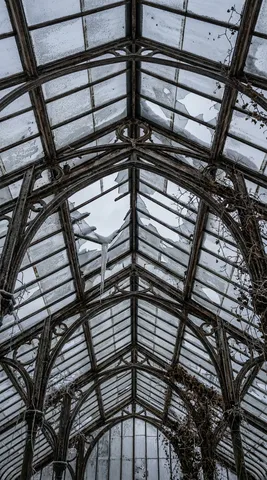 Inside an abandoned Victorian greenhouse in winter, looking straight up at the peaked glass roof. The iron frame creates a gothic arch pattern against the grey sky above. Some glass panels are intact and frosted, some are broken with jagged edges, and snow has entered through the largest break. A single icicle hangs from a broken frame member, catching faint light. Dead vine tendrils cling to the iron ribs. Frost crystals cover every metal surface. The composition is strictly vertical — ground to peak. Shot on Canon R5, 14mm, f/8. Architectural decay photography. Negative: people, summer, bright, text, watermark.