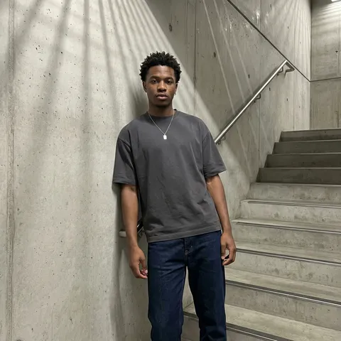 {
  "visual_ontology_schema": {
    "metadata": {
      "aspect_ratio_standard": "4:5 (Vertical - Optimized Feed)",
      "intended_platform_context": "Instagram Grid Post (Permanent)",
      "content_lifecycle": "Live/Raw (Phone Capture)"
    },
    "subject": {
      "description": "A young man standing casually in a modern brutalist stairwell with concrete steps and metal railings, leaning against a textured concrete wall with geometric shadows, captured in a clean street-casual lifestyle portrait with realistic indoor lighting and dramatic directional shadows.",
      "special_rules": {
        "perspective_rules": "Match original composition: subject placed slightly right of frame, stairwell architecture receding diagonally upward into depth, camera at relaxed 3/4 angle not perfectly straight-on; subtle lean; medium-full framing with architectural environment visible",
        "camera_distance_rule": "Camera distance locked to 1.8–2.5 meters (wider framing, more background visible)"
      },
      "demographics": {
        "age": "young adult, early 20s (21-24)",
        "gender_presentation": "Masculine",
        "ethnicity": "Black / African descent"
      },
      "face": {
        "preserve_original": true,
        "expression": "relaxed confident expression with natural ease, calm composed eyes",
        "expression_archetype": "Casual Composed",
        "eyes": {
          "color": "warm brown",
          "details": "ultra-detailed iris texture, crisp limbal ring, realistic catchlights, natural wetline, sharp eyelashes",
          "gaze_rule": "looking directly toward the camera with natural relaxed eye contact"
        },
        "skin": {
          "tone": "deep rich brown skin tone",
          "texture": "realistic skin micro-texture with subtle pores and natural variation (no plastic look), smooth finish",
          "special_detail": "soft highlight on cheekbones with warm indoor reflection"
        },
        "makeup": {
          "style": "Natural Clean Look",
          "details": "natural eyebrows, no eyeliner, minimal grooming, natural lip tone"
        },
        "facial_features": {
          "nose": "soft highlight",
          "lips": "natural brown-red tone, well-defined shape",
          "jawline": "strong and defined"
        }
      },
      "hair": {
        "color": "black",
        "style": "short textured fade with natural volume on top, clean sides",
        "texture": "Textured Curly",
        "finish": "natural matte finish with subtle indoor reflections",
        "length": "Short to Medium"
      },
      "body": {
        "build": "athletic, lean muscular frame",
        "posture": "standing relaxed with shoulders back, leaning subtly against wall, casual confident stance",
        "visible_body_parts": [
          "face",
          "neck",
          "shoulders",
          "arms",
          "upper torso",
          "waist",
          "upper legs"
        ],
        "specific_details": "torso angled slightly, casual relaxed posture with arms naturally positioned"
      },
      "clothing": {
        "overall_style": "street casual / minimalist urban aesthetic",
        "top": {
          "type": "oversized crew neck t-shirt",
          "color": "charcoal gray",
          "details": "relaxed fit, soft cotton texture, clean minimal design",
          "brand_visible": false
        },
        "bottom": {
          "type": "straight-leg jeans",
          "color": "dark indigo",
          "details": "classic fit, tapered at ankle, clean worn look",
          "brand_visible": false
        },
        "fit_and_silhouette": "oversized tee + fitted jeans contrast"
      },
      "pose_and_action": {
        "pose_type": "Standing / Casual Wall Lean",
        "action_description": "standing near the wall in a relaxed pose, leaning naturally with calm confident vibe",
        "hand_placement": "one hand at side, one hand casually touching wall"
      }
    },
    "accessories": {
      "jewelry": {
        "necklace": "simple pendant on thin chain",
        "earrings": "small hoop earring, left ear only",
        "rings": "none"
      },
      "eyewear": {
        "type": "None"
      }
    },
    "background": {
      "setting": "modern brutalist stairwell",
      "location_context": "Indoor (Contemporary Architecture / Stairwell)",
      "wall_color": "raw concrete with geometric shadow patterns and clean architectural lines",
      "floor": "polished concrete floor",
      "elements": [
        [
          "concrete stairwell walls",
          "geometric shadow patterns from overhead lighting",
          "metal handrail visible",
          "stairs receding into architectural depth",
          "minimalist modern ceiling with clean lines"
        ]
      ],
      "depth_and_focus": "sharp subject with moderate background clarity (architecture clearly visible, slight depth separation)",
      "atmosphere": "urban, contemporary, clean architectural vibe"
    },
    "lighting": {
      "primary_source": "overhead architectural lighting with dramatic directional quality",
      "quality": "moderately hard light creating geometric wall shadows, clean contemporary contrast",
      "direction": "top-front overhead light with side directional elements",
      "color_temperature": "neutral indoor (4000-5000K)",
      "mood": "urban and contemporary, realistic indoor architectural lighting"
    },
    "technical_acquisition": {
      "device_class": "Smartphone (Flagship)",
      "specific_hardware_guess": "Apple iPhone 14/15 Pro",
      "camera_mode": "Standard Wide",
      "camera_distance": "1.8–2.5 meters",
      "camera_angle": "eye-level with subtle tilt, mild 3/4 perspective (not perfectly straight-on)",
      "framing": "Medium Full Shot (MFS) / 3/4 Body",
      "lens_character": "natural phone wide-lens look (24–28mm equivalent), subtle perspective depth",
      "digital_artifacts": [
        "clean phone sharpening",
        "mild indoor noise reduction",
        "crisp texture on wall and outfit"
      ]
    },
    "aesthetic_and_style": {
      "overall_archetype": "Urban Contemporary Lifestyle (Architectural Portrait)",
      "color_palette": {
        "dominant_colors": [
          "raw concrete gray",
          "charcoal gray top",
          "dark indigo jeans",
          "deep brown skin tone",
          "black hair",
          "geometric shadow patterns"
        ],
        "color_harmony": "neutral contemporary urban",
        "saturation_level": "natural"
      },
      "visual_references": [
        "brutalist architecture street portraits",
        "contemporary minimalist lifestyle aesthetic",
        "urban candid phone photography"
      ]
    },
    "post_processing": {
      "filter_aesthetic": "Clean Contemporary Editorial",
      "editing_intensity": "Light to Moderate enhancement",
      "specific_edits": {
        "color_grading": "neutral grays, balanced warm skin tones, preserved architectural detail",
        "skin_retouching": "light smoothing preserving natural texture",
        "background_manipulation": [
          "slight contrast control to maintain shadow definition"
        ]
      }
    },
    "contextual_intent": {
      "primary_purpose": "street lifestyle branding / contemporary portrait post",
      "target_audience": "Public followers",
      "engagement_strategy": [
        "Urban vibe",
        "Clean contemporary aesthetic",
        "Confident minimalism"
      ]
    },
    "negative_prompt": "different face identity, light hair, light eyes, low quality, blurry, pixelation, jpeg artifacts, compression artifacts, banding, low resolution, heavy noise, overexposed face, harsh flash, plastic skin, over-smoothed skin, distorted face, deformed anatomy, extra fingers, bad hands, watermark, text, logo, UI overlay, nudity, explicit content"
  }
}